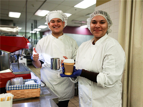Two culinary students in a professional kitchen, one giving a thumbs up while holding a metal pitcher and the other holding a paper cup, both wearing white chef jackets and hats, with a red espresso machine in the background.