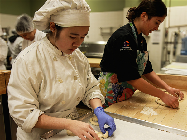 Focused culinary students working in a commercial kitchen, one kneading dough on a wooden surface and another in a black chef coat with a colorful apron shaping dough in the background.