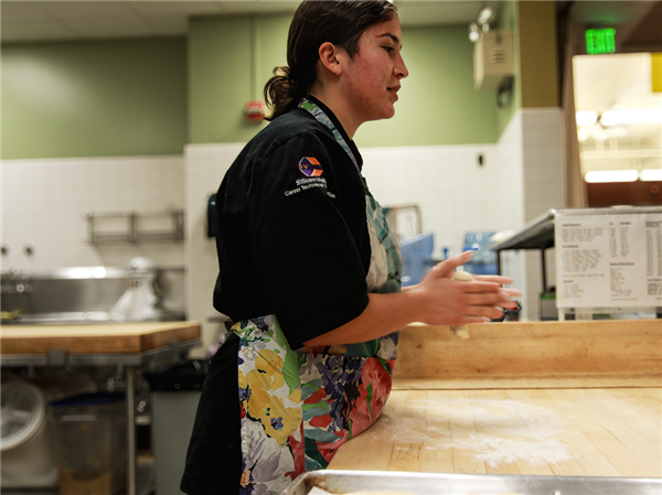 A student in a black chef jacket and colorful apron attentively shaping dough on a wooden counter in a commercial kitchen, with a focus on the hands and the dough.