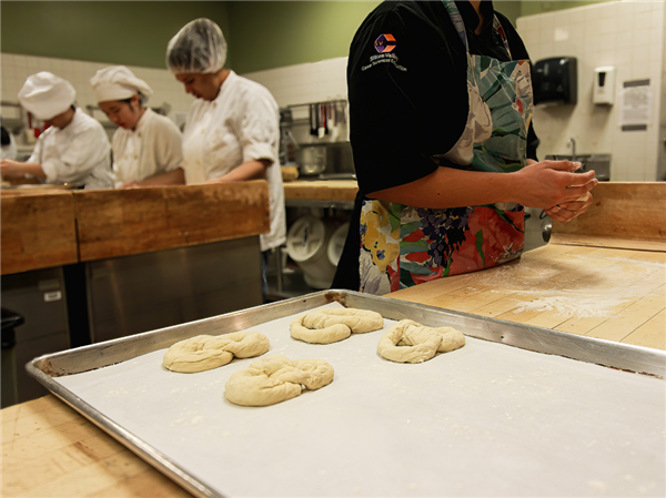 A bustling culinary classroom with students in white chef attire working on various tasks; one in the foreground is shaping dough on a baking sheet on a wooden bench.
