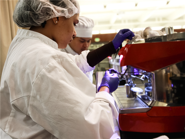 wo students operating a red commercial espresso machine in a professional kitchen, wearing white chef attire and hairnets, focusing on the task with a sense of concentration.