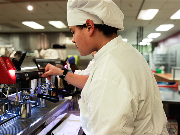 A culinary student in a white chef hat and jacket is intently operating an espresso machine, with buttons and a steaming wand, in a commercial kitchen setting.