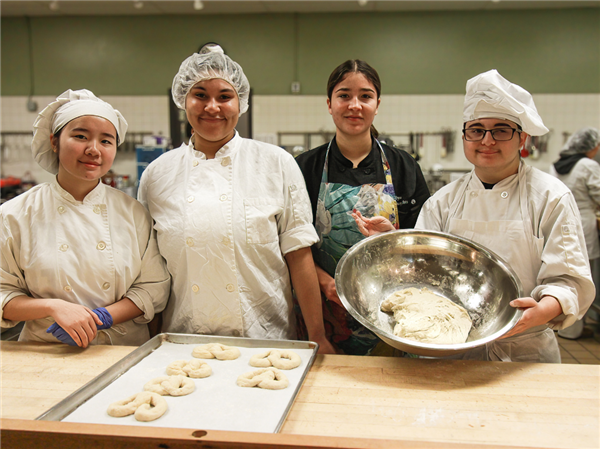 Four culinary students posing in a kitchen, three in white chef jackets and one in a black chef coat with a colorful apron, with a bowl of dough and baking trays in front of them.